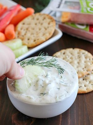 A cucumber being dipped into a creamy Cucumber Dill Dip with a background of crackers and veggies + 43 Healthy Snack Ideas