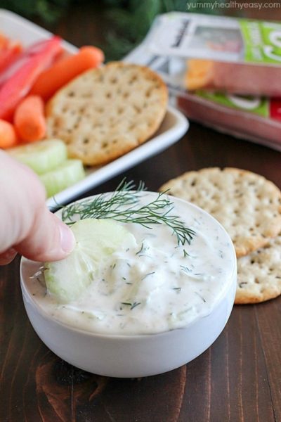 A cucumber being dipped into a creamy Cucumber Dill Dip with a background of crackers and veggies.