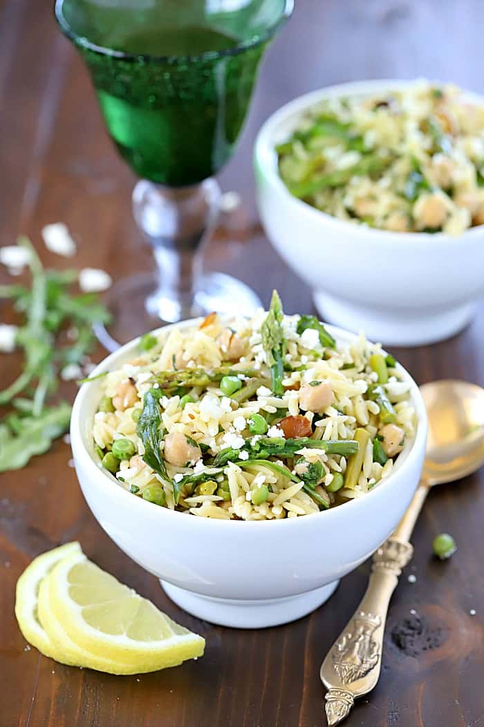 Two bowls filled with a beautiful Spring Salad filled with orzo, asparagus, toasted almonds, arugula, feta cheese and peas with a vinaigrette.