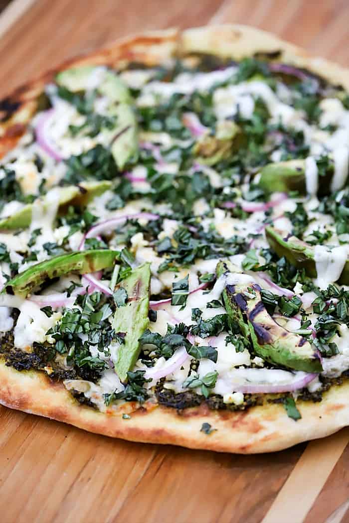Close up view of cooked grilled pizza topped with pesto, cheeses, red onion, grilled avocado and basil.
