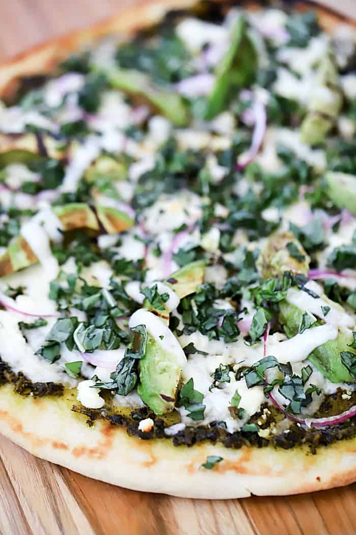 Close up view of cooked grilled pizza topped with pesto, cheeses, red onion, grilled avocado and basil.