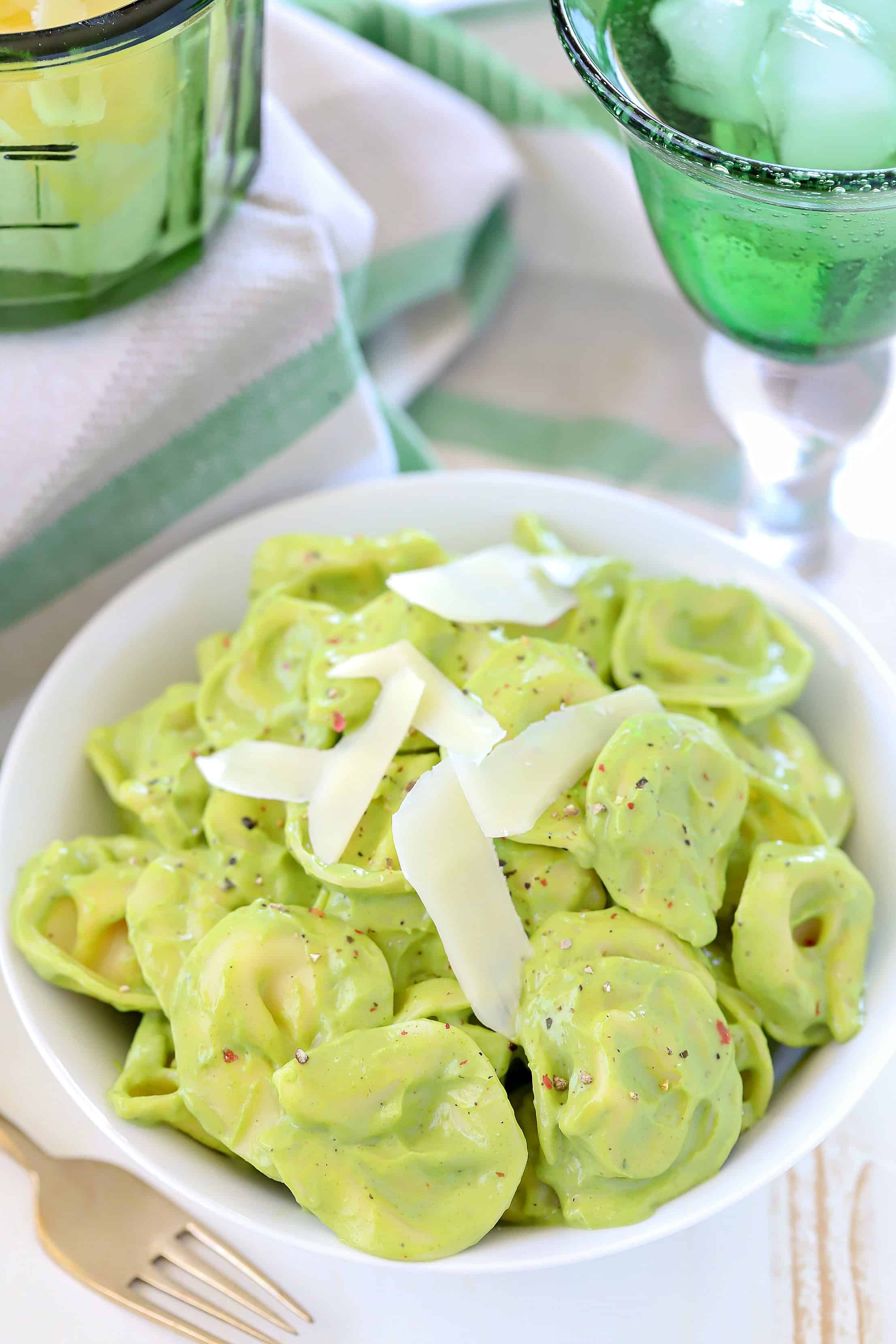 White bowl filled with tortellini coated in an avocado cream sauce with shaved parmesan on top. 